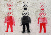 Se ven tres muñecos, que parecen de playmobil,  pintados en una pared, con un spray usando la mima plantilla,   cada uno con una bombilla encima. Los dos de los lados en rojo, y el del centro en negro. 
