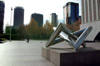 Monumento ubicado en una gran plaza. El monumento es un tubo de metal que está anudado. Al fondo se ven edificios de oficinas. Zona empresarial de Madrid.