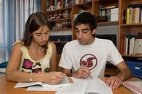 Elena explicando contabilidad a Luis. Fotografía en la que aparece Elena a la izquierda, señalando algo en un libro, y Luís, mirando atentamente. En la fotografía Elena está explicando contabilidad a Luis, en una mesa con varios libros y documentos.