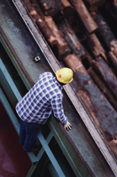 La fotografía muestra, desde una posición de cámara muy alta, a un operario con un casco de protección amarillo asomándose por encima de una estructura metálica observando unos troncos apilados.