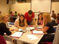 Reunión en una sala amplia de varios grupos de trabajo. En primer plano uno de ellos,  sentados alrededor de una mesa y hablando sobre el trabajo. Cuatro mujeres están sentadas, una hablando y las otras tres mirándola con atención. El quinto miembro del equipo es un hombre, que está de pie, con los brazos apoyados sobre la mesa, y mirando fijamente un documento que hay encima de esa mesa.