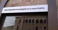 Letrero identificativo del Centro Internacional de Investigación de la Lengua Española. Debajo del mismo hay una puerta con cristales de espejo sobre la que se refleja un edificio antiguo de fachada en piedra, y una arcada en el piso superior. 