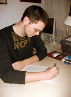 Imagen de Ricardo de perfil sentado y escribiendo sobre una mesa. Viste camiseta negra de manga larga y lleva gafas.