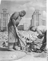 Fotografía en blanco y negro que muestra dos trabajadores de la construcción. El trabajador de la izquierda cubre su cabeza con una especie de boina e está manejando un martillo neumático. El de la derecha está retirando el escombro.