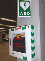 Dispositivo desfibrilador externo automático en una vitrina en un lugar público, (un aeropuerto) con la correspondiente señalización de su ubicación colocada encima del mismo, un cartel verde, con borde blanco, y un corazón blanco partido por un rayo, y el texto AED. (Automatic External Defibrillator, o Desfibrilador Externo Automático).