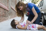 Claudia está arrodillada junto a una niña tumbada en el suelo. Está comprobando la consciencia de la niña.