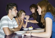 Cuatro jóvenes sentados en una mesa de restaurante. Chico joven sentado en un restaurante frente a una muchacha. A su lado otra chica se lleva las manos al cuello. Frente a ella, una chica de pelo oscuro se inclina a ayudarla desde el otro lado de la mesa.