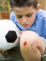 Fotografía de un niño de unos siete años con una pelota de fútbol. Está agachado mirándose una pequeña herida en la rodilla. Hay restos de hierba alrededor de la herida por lo que parece producida por una caída mientras jugaba.