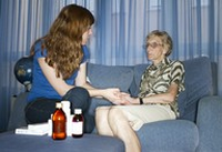 Una mujer joven está sentada en un sofá en casa de su abuela. Sujeta la mano de la abuela que permanece recostada en el sofá. En una mesa se ven diversos medicamentos.