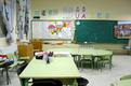 Se ve un aula de infantil, con la mesa del profesor, con la pizarra y un tablón de anuncios al fondo, y con mesas y sillas para los niños. Muchas letras de colores y dibujos infantiles colgados de la pared. 