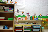 Rincón de un alua de infantil en el que se ven los armarios donde se guardan los materiales parra jugar, pintar, hacer manualidades, etc. Cada recipiente etiquetado con el nombre de un niño o niña, y de fondo, la pared empapelada y con dibujos y recortes infantiles pegados. 