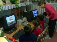 Fotograma de un vídeo en el que se puede ver a una maestra en clase explicando algo a unos niños. Los niños están sentados en frente del ordenador, mientras que la maestra esta justo detrás de ellos observando lo que hacen. Los niños están usando el ordenador con algún tipo de software.