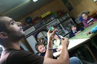 Fotografía en la que se puede ver a un maestro de educación infantil explicando algo a un grupo de niños. Los niños están sentados alrededor de la mesa, mirando atentamente al maestro. El maestro aparece en primer plano, mirando a los alumnos, y entre sus manos tiene un lápiz. Al fondo de la fotografía se puede apreciar una estantería repleta de libros y otros objetos. 