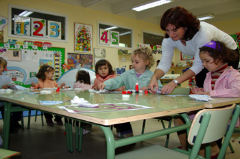 Fotografía que muestra a una educadora dando clases y explicando en un aula de un centro infantil. En la fotografía aparece la educadora mostrando como se realiza una tarea, mientras la niña mira atentamente. La clase versa sobre manualidades.