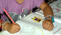  Fotografía de una niña, o un niño, al que no se le ve la cara, que está realizando sus tareas escolares. Concretamente está dibujando sobre un cuadernillo que ya trae los dibujos hechos con líneas discontinuas.