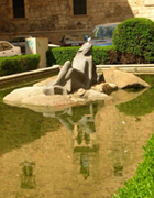 Escultura de una mujer tomando el sol sobre unas piedras, en medio de un estanque en el que se refleja la escultura y el edificio del fondo.