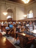 Eva estudiando delante del ordenador en una sala de una biblioteca  que parece ser de un edificio histórico, con mesas de madera,  paredes de piedra, y artesonado en el techo. Todas las mesas están llenas de ordenadores de pantalla plana, que dan un toque de modernidad en un ambiente tradicional.