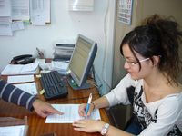 Imagen con un plano medio de Blanca, sentada en una mesa, firmando papeles, mientras se ve la mano de alguien que le va indicando con el dedo algún detalle del documento. ”