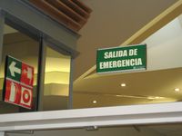 Detalle de la señalización de emergencia en un edificio. Se ven varias señales, que indican distintos elementos de seguridad  contra incendios, pero el foco de la imagen está en el letrero de fondo verde y letras blancas con el texto SALIDA DE EMERGENCIA.