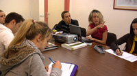 Imagen de una reunión de empresa en la que aparecen varias personas sentadas alrededor de una mesa. Todos están tomando apuntes a excepción de una mujer que, junto a un proyector, explica algo a sus compañeros.